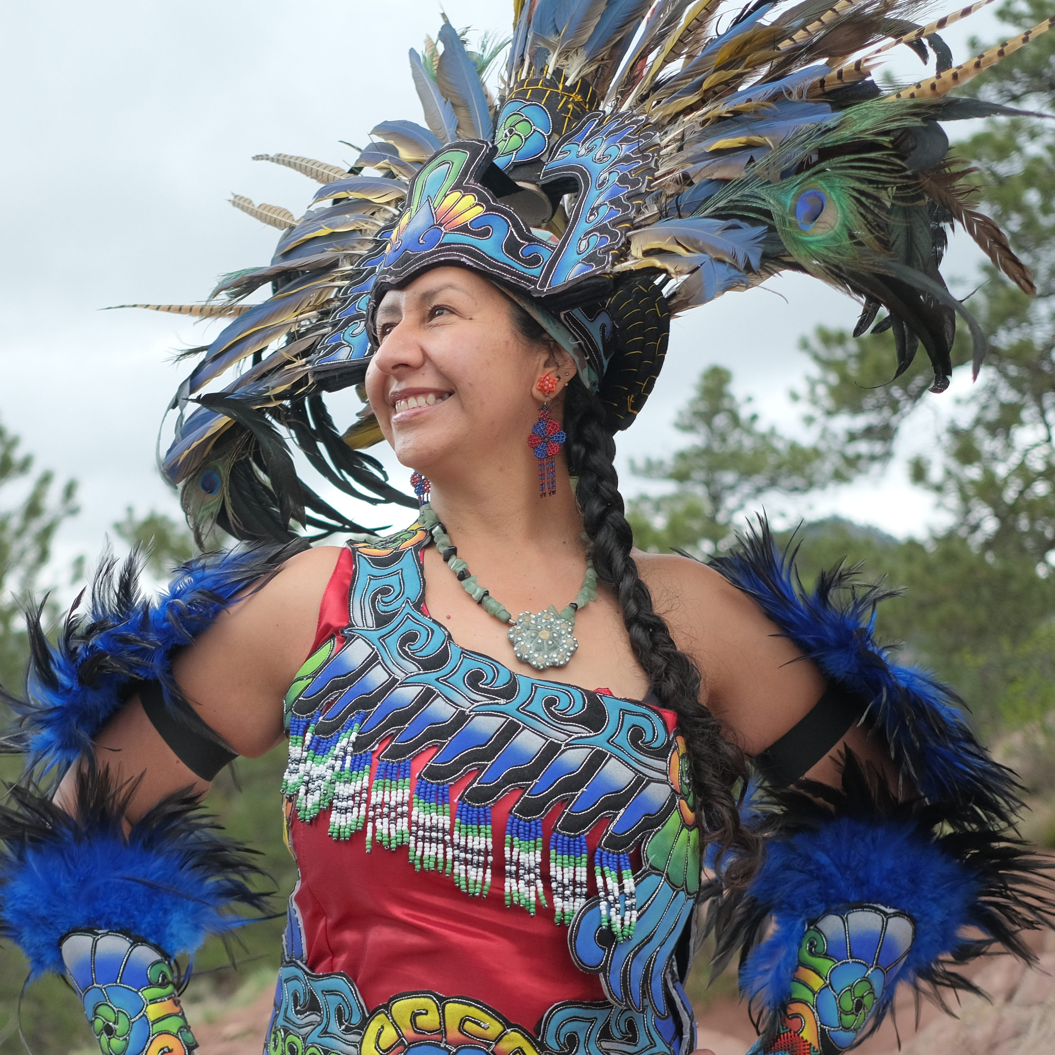 Andrea smiling and looking into the distance wearing bright Azteca dance regalia and abright feather headpiece